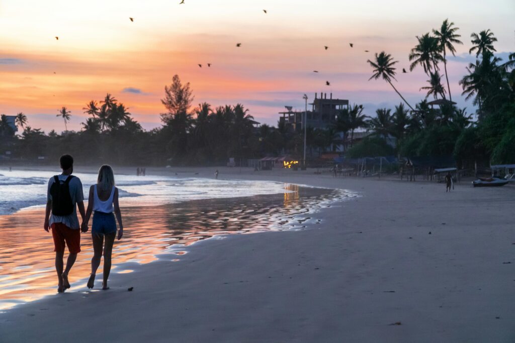 Back view of full body unrecognizable couple in casual summer clothes holding hands and strolling along sandy seashore in tropical resort at sunset with picturesque sky