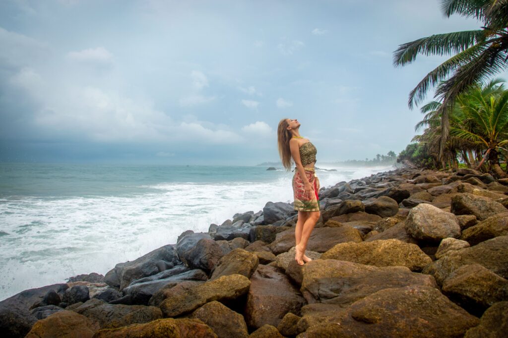 Woman standing on rocks by the ocean, enjoying the scenic view under a cloudy sky.