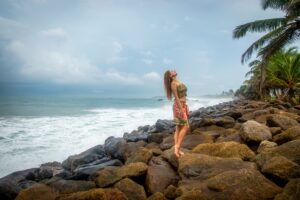 Woman standing on rocks by the ocean, enjoying the scenic view under a cloudy sky.